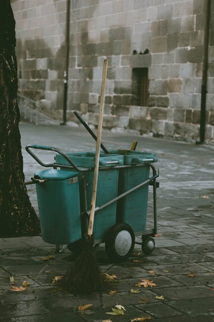 A man with short dark hair, dressed in a black T-shirt, is seen collecting litter in a white garbage bag with both hands while leaning over a metal public trash bin on a paved sidewalk. The trash bin