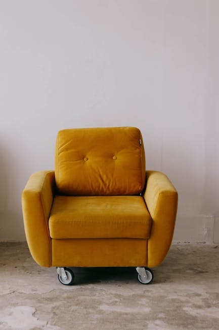 A mustard yellow armchair with a cushioned backrest featuring button details, placed on four small caster wheels. The armchair is positioned against a plain white wall in a room with a light-colored, 