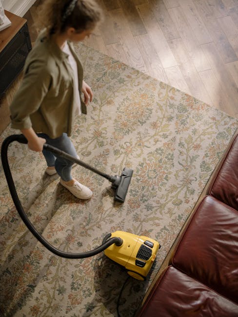 A professional cleaning specialist dressed in a white protective suit, mask, gloves, and shoe covers is conducting deep cleaning on a beige plush carpet in a modern living room. The room features a ta